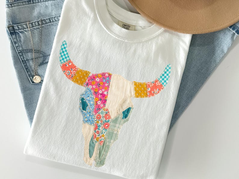 May include: White t-shirt featuring a colorful patchwork bull skull design. The skull is made of various patterned fabrics in shades of pink, blue, yellow, and green. The shirt is paired with blue jeans and a tan hat.