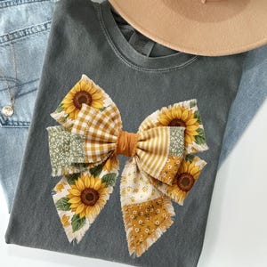 May include: A gray t-shirt featuring a large bow made of various patterned fabrics, including sunflowers, gingham, and floral prints. The bow is centered on the shirt, with a tan hat and blue jeans in the background. A gold necklace is visible.