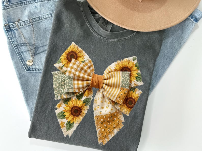 May include: A gray t-shirt featuring a large bow made of various patterned fabrics, including sunflowers, gingham, and floral prints. The bow is centered on the shirt, with a tan hat and blue jeans in the background. A gold necklace is visible.