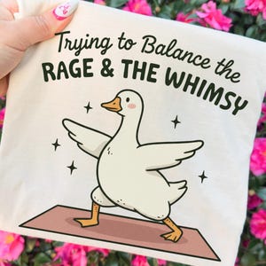 May include: A cream-colored tote bag featuring a cartoon goose in a yoga pose on a brown mat. The text "Trying to Balance the RAGE & THE WHIMSY" is printed above the goose in black. Four small black stars surround the goose.