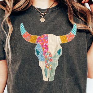 May include: Dark gray t-shirt featuring a colorful patchwork bull skull design. The skull is adorned with various floral and patterned fabrics in shades of pink, blue, yellow, and green. The shirt has a classic crew neck.