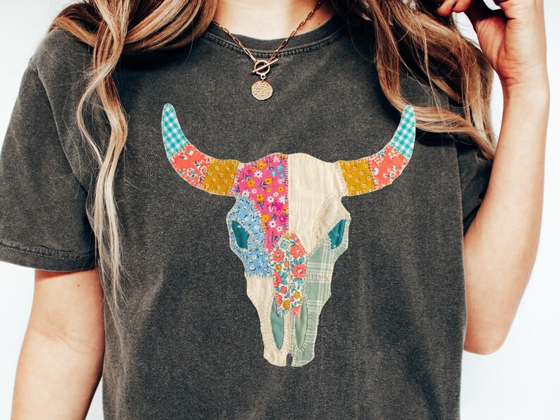 May include: Dark gray t-shirt featuring a colorful patchwork bull skull design. The skull is adorned with various floral and patterned fabrics in shades of pink, blue, yellow, and green. The shirt has a classic crew neck.