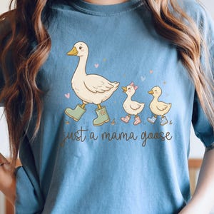 May include: Blue t-shirt featuring a whimsical illustration of a mother goose and two goslings, all wearing boots. The text "just a mama goose" is printed below the image. The design includes hearts and grass.