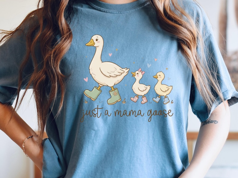 May include: Blue t-shirt featuring a whimsical illustration of a mother goose and two goslings, all wearing boots. The text "just a mama goose" is printed below the image. The design includes hearts and grass.