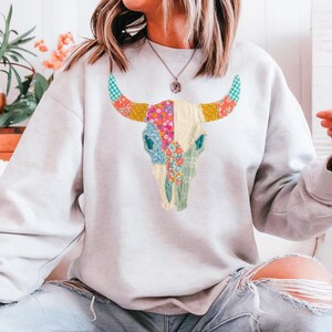May include: A light gray sweatshirt featuring a colorful bull skull design. The skull is decorated with floral and patterned patches in shades of pink, blue, yellow, and green. The horns have a blue and white checkered pattern.