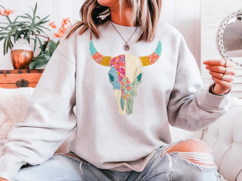 May include: A light gray sweatshirt featuring a colorful bull skull design. The skull is decorated with floral and patterned patches in shades of pink, blue, yellow, and green. The horns have a blue and white checkered pattern.