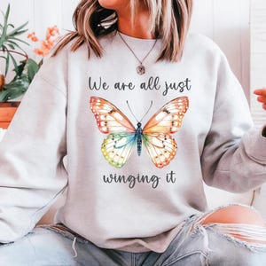 May include: Light gray sweatshirt featuring a colorful butterfly design with orange, yellow, blue, and green wings. The text "We are all just winging it" is above and below the butterfly illustration.