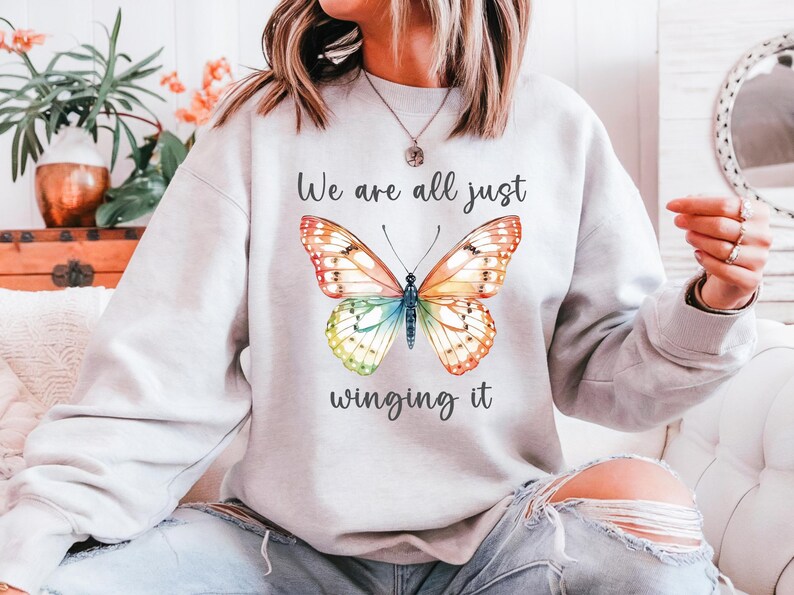 May include: Light gray sweatshirt featuring a colorful butterfly design with orange, yellow, blue, and green wings. The text "We are all just winging it" is above and below the butterfly illustration.
