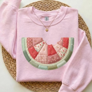 May include: A light pink sweatshirt featuring a patchwork watermelon slice design. The watermelon slice is made of various fabrics in shades of red, pink, and green. A gold necklace with a heart pendant is also visible.
