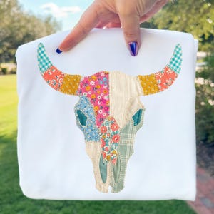 Könnte beinhalten: Weißes Sweatshirt mit einem Patchwork-Design eines Bullenschädels. Der Schädel ist mit floralen, karierten und gemusterten Stoffen in Rosa, Gelb, Blau und Grün verziert. Die Hörner haben ein türkis-weißes Karomuster.
