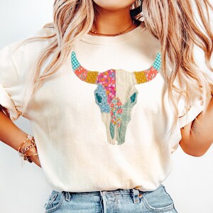 May include: Cream-colored t-shirt featuring a colorful patchwork bull skull design. The skull is adorned with floral, plaid, and patterned fabric in shades of pink, blue, yellow, and green. The shirt has short sleeves and a relaxed fit.