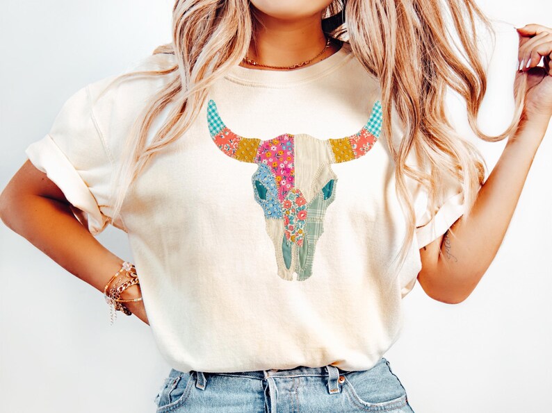May include: Cream-colored t-shirt featuring a colorful patchwork bull skull design. The skull is adorned with floral, plaid, and patterned fabric in shades of pink, blue, yellow, and green. The shirt has short sleeves and a relaxed fit.