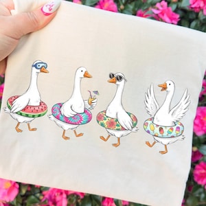May include: A cream-colored tote bag featuring four cartoon geese, each wearing a different style of inflatable ring and sunglasses. One goose has wings spread. The rings have watermelon, tie-dye, floral, and popsicle designs.