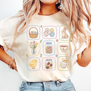 May include: Cream-colored t-shirt featuring a grid of Easter-themed illustrations. The design includes a basket of eggs, decorated eggs, a bunny, flowers, jelly beans in a jar, a teacup, a chick, and a decorated egg.