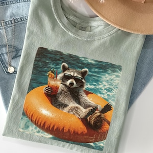 May include: A light green t-shirt featuring a raccoon wearing sunglasses and floating on an orange inflatable ring in a pool. The raccoon is holding a cocktail. The shirt is laid on top of blue jeans and a tan hat.
