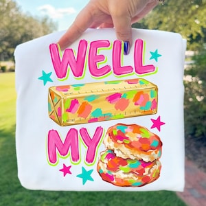 May include: A white t-shirt featuring a graphic print with the text WELL MY in bright pink. Below the text is a gold stick of butter with colourful paint-like splatters and a stack of two biscuits with similar colourful splatters and teal stars.