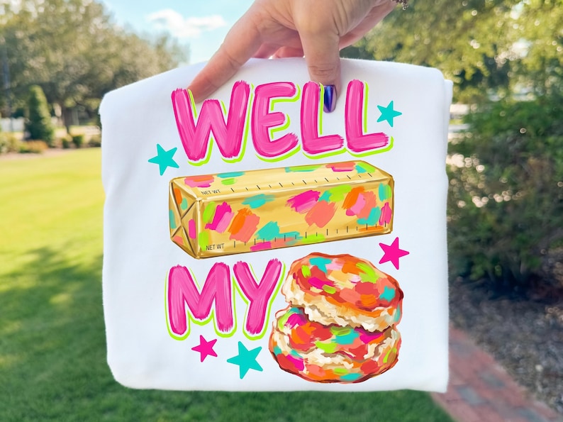 May include: A white t-shirt featuring a graphic print with the text WELL MY in bright pink. Below the text is a gold stick of butter with colourful paint-like splatters and a stack of two biscuits with similar colourful splatters and teal stars.