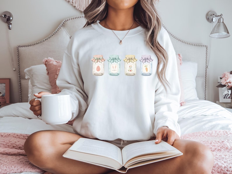 May include: A white sweatshirt featuring four jars with Easter-themed designs. Each jar has a different pastel-colored lid and contains an egg, bunny ears, flowers, and a carrot. The sweatshirt is worn by a person holding a white mug and a book.