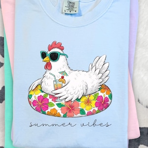 May include: Pale blue t-shirt featuring a cartoon chicken in sunglasses, relaxing on a floral inflatable ring. The words "summer vibes" are written below. The shirt is folded with other pastel coloured shirts.