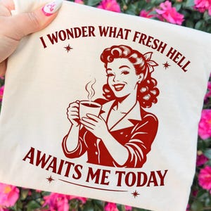 May include: Off-white tote bag with a retro illustration in red. The image features a woman holding a coffee cup, with the text "I WONDER WHAT FRESH HELL AWAITS ME TODAY". The bag is held in front of a background of pink flowers.