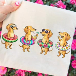 Pode incluir: Uma sacola bege com uma ilustração colorida de quatro dachshunds aproveitando o verão. Cada cão usa um anel inflável diferente, incluindo um design de melancia, tie-dye, floral e picolé. Um cão usa óculos de natação, outro óculos de sol.