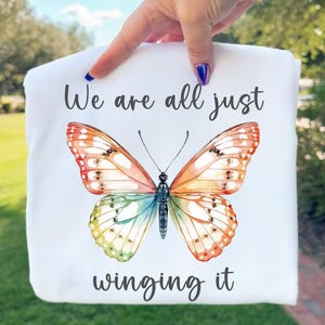 May include: White t-shirt featuring a colorful butterfly illustration with orange, yellow, green, and blue wings. The text "We are all just winging it" is written above and below the butterfly in a script font.