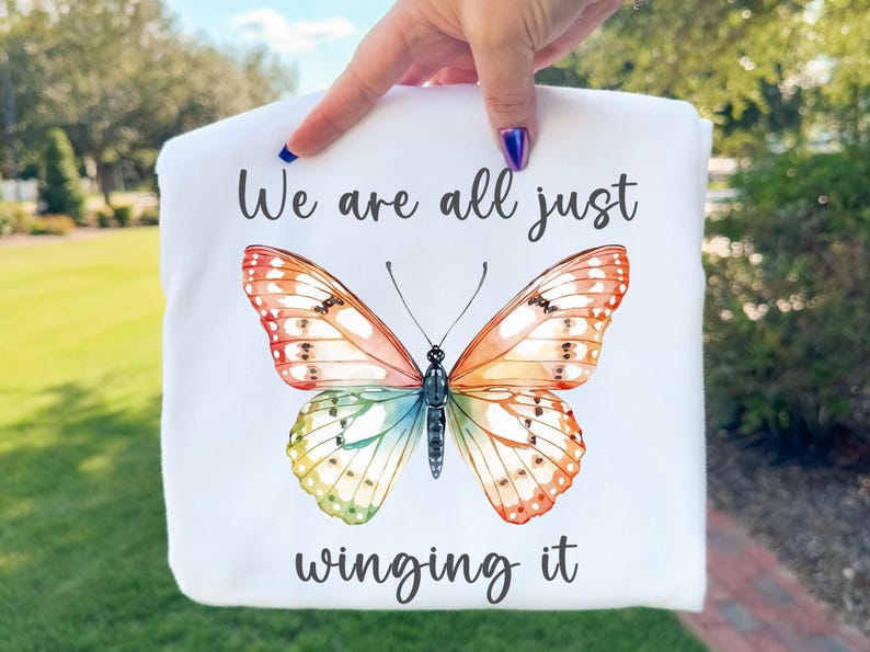 May include: White t-shirt featuring a colorful butterfly illustration with orange, yellow, green, and blue wings. The text "We are all just winging it" is written above and below the butterfly in a script font.
