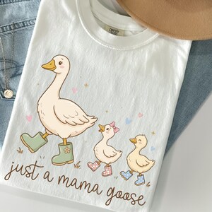 May include: White t-shirt with a whimsical illustration of a mama goose and two goslings, all wearing boots. The text "just a mama goose" is written in a cursive font. The shirt is laid on top of blue jeans and a tan hat.