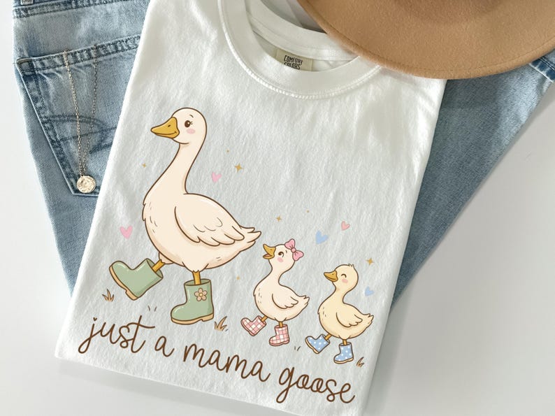 May include: White t-shirt with a whimsical illustration of a mama goose and two goslings, all wearing boots. The text "just a mama goose" is written in a cursive font. The shirt is laid on top of blue jeans and a tan hat.