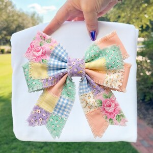 Pu&ograve; includere: Un grande fiocco patchwork colorato con bordi sfilacciati. Il fiocco presenta vari motivi floreali e a quadretti in tonalit&agrave; pastello di rosa, blu, giallo e verde. Il fiocco &egrave; tenuto su uno sfondo bianco.
