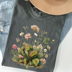 May include: A dark grey t-shirt with a botanical illustration of a Venus flytrap and wildflowers in pink, white, and green. The shirt is shown with light blue jeans, a gold necklace, and a beige hat.