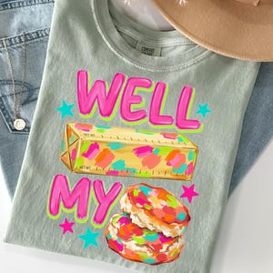 May include: A sage green t-shirt featuring the text WELL MY in bright pink with a neon green outline. Below the text is a yellow stick of butter with colourful paint-like splotches and a biscuit with similar colourful splotches, accented by stars.