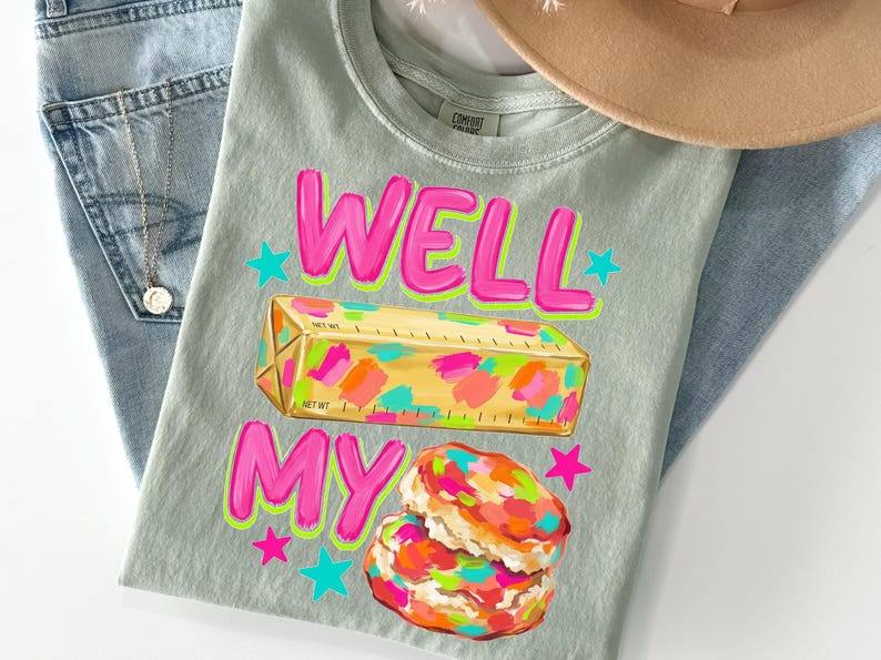 May include: A sage green t-shirt featuring the text WELL MY in bright pink with a neon green outline. Below the text is a yellow stick of butter with colourful paint-like splotches and a biscuit with similar colourful splotches, accented by stars.