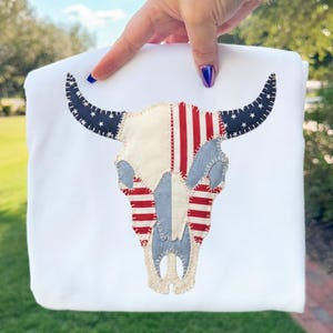 Op de afbeelding: Witte sweatshirt met een stierenkopontwerp. De schedel is versierd met rood-witte strepen, blauwe denim en blauwe sterren, die lijken op de Amerikaanse vlag. De hoorns zijn marineblauw met witte sterren.