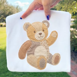 May include: White fabric featuring a patchwork teddy bear design. The bear is in shades of brown and tan, with a smiling face and an outstretched arm. Likely for a baby blanket or nursery decor.