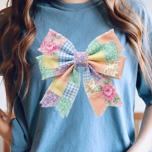 Pu&ograve; includere: T-shirt azzurra con un grande design a fiocco patchwork. Il fiocco incorpora vari colori pastello e motivi floreali, tra cui rose rosa, quadretti vichy e colori uniti. Il fiocco &egrave; il punto focale della maglietta.
