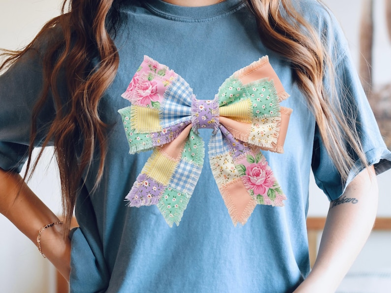 Pu&ograve; includere: T-shirt azzurra con un grande design a fiocco patchwork. Il fiocco incorpora vari colori pastello e motivi floreali, tra cui rose rosa, quadretti vichy e colori uniti. Il fiocco &egrave; il punto focale della maglietta.