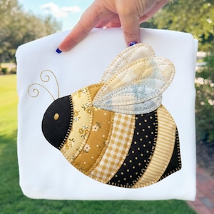 Könnte beinhalten: Ein weißes Stoffstück mit einer großen, applizierten Biene. Die Biene hat einen schwarzen Kopf, einen gelb-schwarz gestreiften Körper und gemusterte Flügel in Gelb-, Blau- und Weißtönen. Die Fühler der Biene sind mit Goldfaden bestickt.