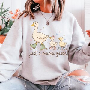 May include: A light gray sweatshirt features a whimsical illustration of a mother goose and her goslings. The mama goose wears green boots, and the text "just a mama goose" is below the illustration. The goslings wear boots and a bow.