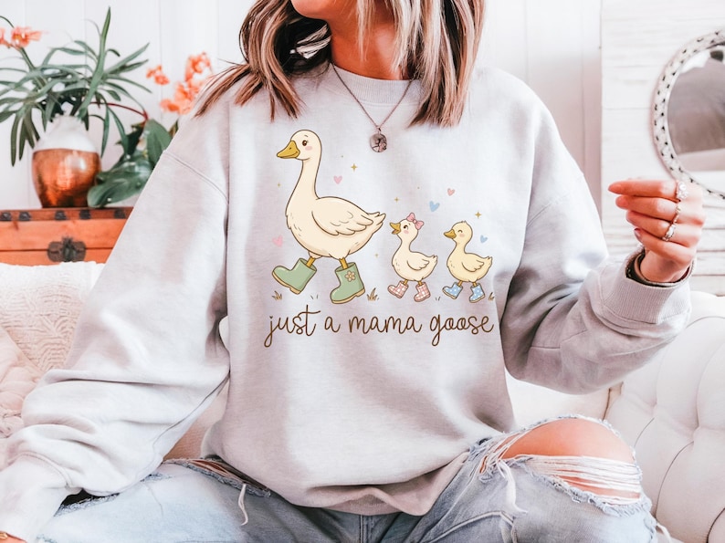 May include: A light gray sweatshirt features a whimsical illustration of a mother goose and her goslings. The mama goose wears green boots, and the text "just a mama goose" is below the illustration. The goslings wear boots and a bow.