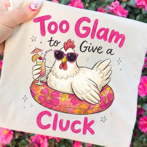 May include: A cream-colored tote bag featuring a cartoon chicken wearing sunglasses and floating on a floral inner tube, holding a cocktail. The text "Too Glam to Give a Cluck" is written in pink.