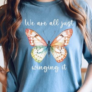 May include: A blue t-shirt featuring a colorful butterfly design. The butterfly has orange, yellow, and blue wings. The text "We are all just winging it" is written in white script above and below the butterfly.