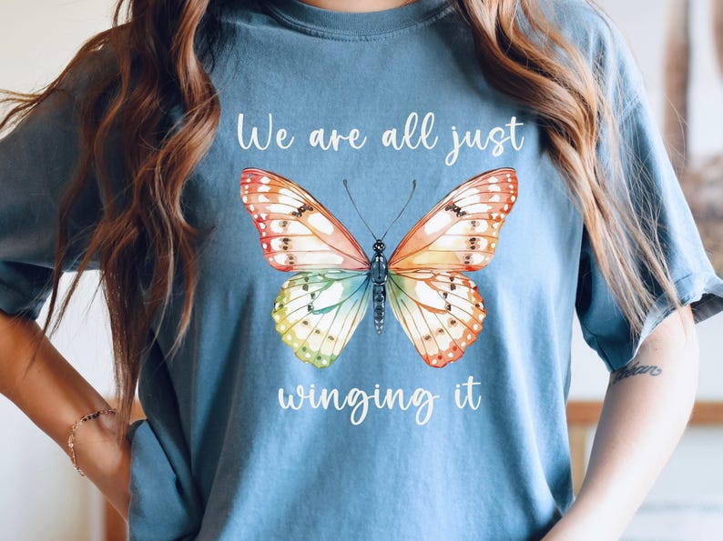 May include: A blue t-shirt featuring a colorful butterfly design. The butterfly has orange, yellow, and blue wings. The text "We are all just winging it" is written in white script above and below the butterfly.
