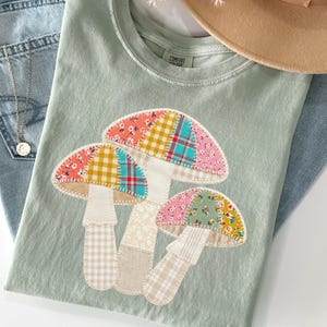 May include: A light green t-shirt featuring a colorful mushroom design made of patchwork fabric. The mushrooms have patterned caps in pink, yellow, and blue. The shirt is paired with blue jeans and a tan hat.