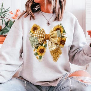 May include: A light gray sweatshirt featuring a large bow design. The bow is composed of various patterned fabrics, including sunflowers, gingham, and floral prints in yellow, green, and white. The sweatshirt is casual wear.