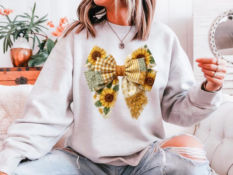 May include: A light gray sweatshirt featuring a large bow design. The bow is composed of various patterned fabrics, including sunflowers, gingham, and floral prints in yellow, green, and white. The sweatshirt is casual wear.