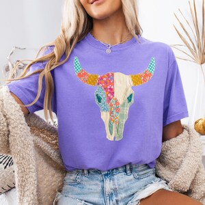 May include: A purple t-shirt featuring a colorful patchwork bull skull design. The skull is cream-colored with floral and patterned accents in pink, green, and yellow. The horns have a checkered pattern. The shirt is a comfortable fit.