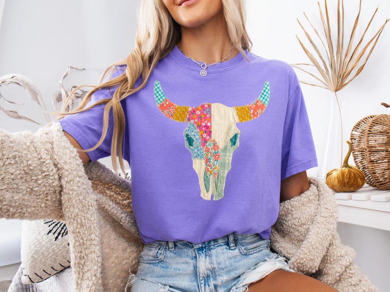 May include: A purple t-shirt featuring a colorful patchwork bull skull design. The skull is cream-colored with floral and patterned accents in pink, green, and yellow. The horns have a checkered pattern. The shirt is a comfortable fit.
