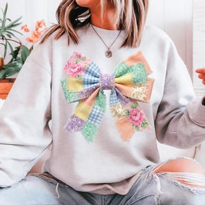 Pu&ograve; includere: Una felpa grigio chiaro con un grande design a fiocco patchwork colorato. L'arco incorpora motivi floreali, quadretti e colori pastello a tinta unita. L'arco &egrave; il punto focale della felpa, creando un look stravagante e artistico.