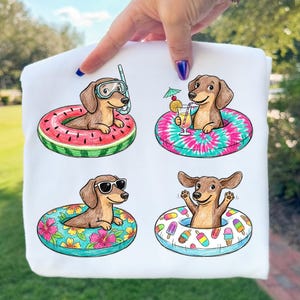 Pode incluir: Camisola branca com quatro ilustrações de cachorros salsicha em boias coloridas. Um cão usa um snorkel, outro bebe um coquetel, um terceiro usa óculos de sol e o último tem as patas levantadas. As boias têm desenhos de melancia, tie-dye, floral e picolé.